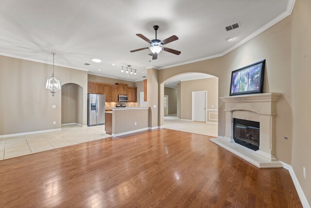 unfurnished living room featuring visible vents, light wood finished floors, arched walkways, a glass covered fireplace, and ceiling fan with notable chandelier