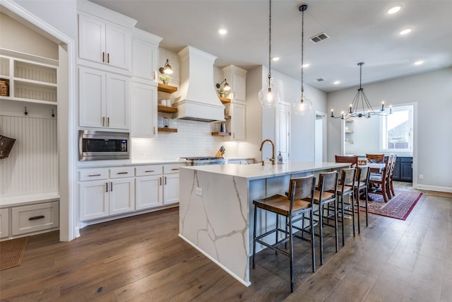 kitchen with visible vents, a center island with sink, custom exhaust hood, open shelves, and stainless steel microwave