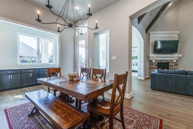 dining room with light wood-type flooring, arched walkways, a stone fireplace, a chandelier, and vaulted ceiling with beams