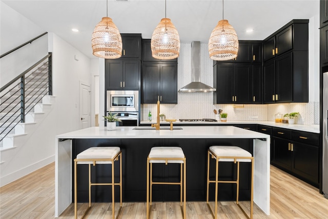 kitchen with light countertops, light wood-style floors, stainless steel appliances, wall chimney exhaust hood, and dark cabinets