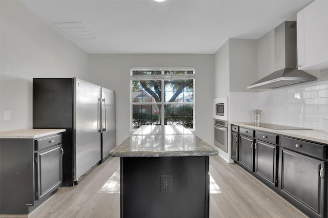 kitchen featuring a kitchen island, appliances with stainless steel finishes, wall chimney exhaust hood, light wood-type flooring, and backsplash