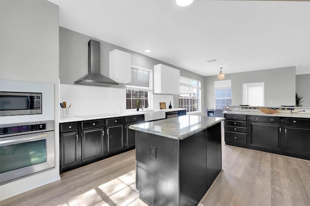 kitchen with dark cabinetry, a kitchen island, stainless steel appliances, light wood-style floors, and wall chimney exhaust hood