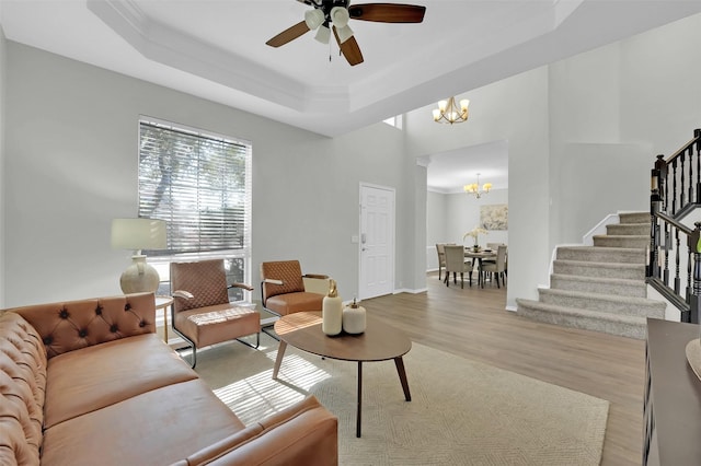 living room with stairs, crown molding, a raised ceiling, ceiling fan with notable chandelier, and light wood-type flooring