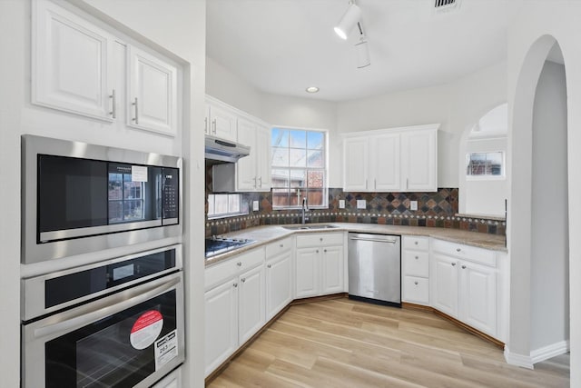 kitchen with light wood-style flooring, a sink, under cabinet range hood, tasteful backsplash, and appliances with stainless steel finishes