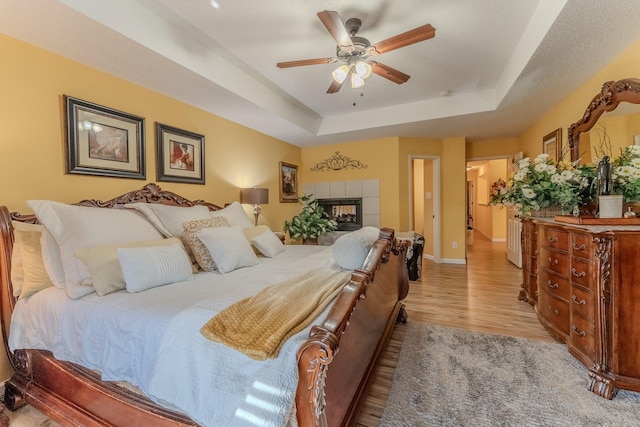 bedroom with ceiling fan, baseboards, a tile fireplace, wood finished floors, and a raised ceiling