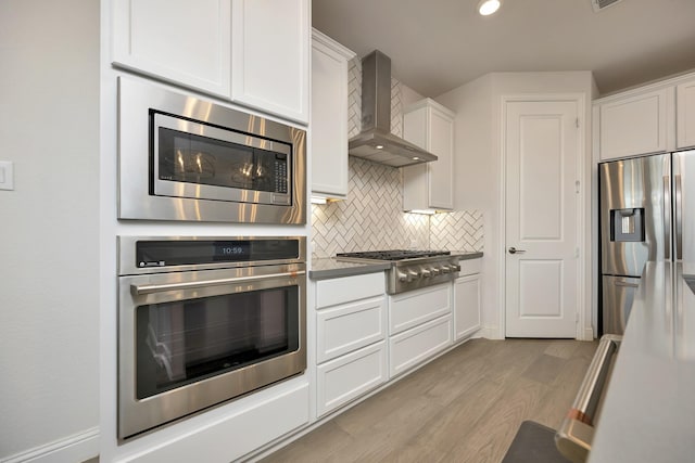 kitchen with decorative backsplash, light wood-style floors, white cabinets, stainless steel appliances, and wall chimney exhaust hood