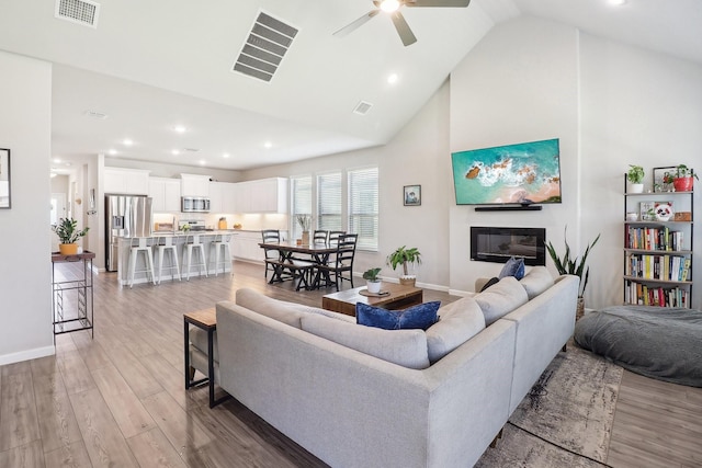 living room featuring a glass covered fireplace, visible vents, high vaulted ceiling, and light wood-style flooring