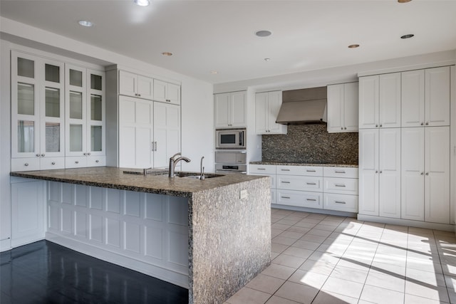 kitchen featuring stainless steel microwave, decorative backsplash, dark stone countertops, custom exhaust hood, and a sink