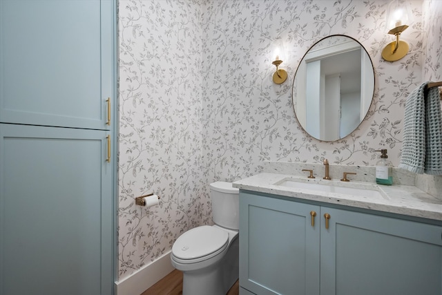 half bath with baseboards, toilet, vanity, and wallpapered walls