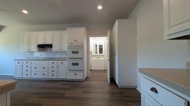 kitchen featuring under cabinet range hood, backsplash, dark wood-style floors, and white double oven