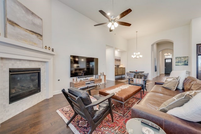 living room featuring wood finished floors, baseboards, arched walkways, a tile fireplace, and ceiling fan with notable chandelier