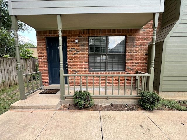 doorway to property featuring brick siding, a porch, and fence