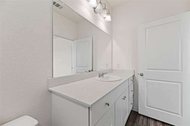 half bath featuring vanity, wood finished floors, visible vents, toilet, and a textured wall