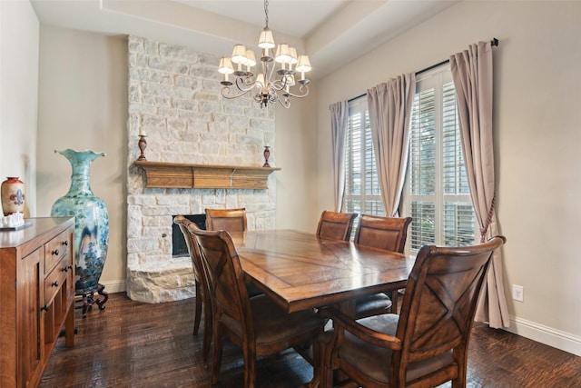 dining space with baseboards, dark wood finished floors, a stone fireplace, a raised ceiling, and a chandelier