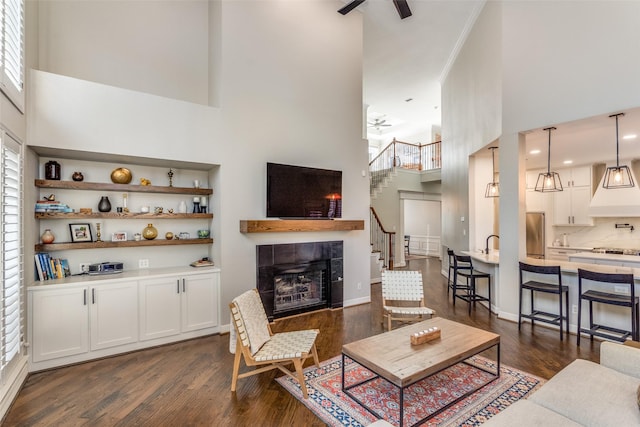 living room featuring ceiling fan, stairway, a fireplace, a towering ceiling, and dark wood-style flooring