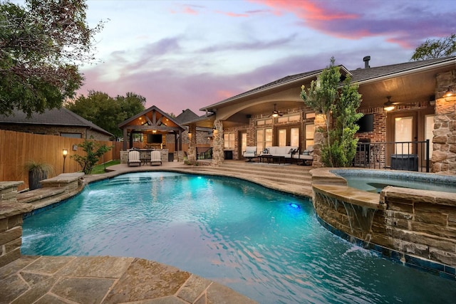 pool at dusk featuring fence, an outdoor living space with a fireplace, a pool with connected hot tub, ceiling fan, and a patio area