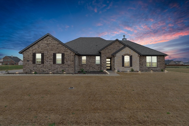 french country home featuring stone siding, a lawn, roof with shingles, and brick siding