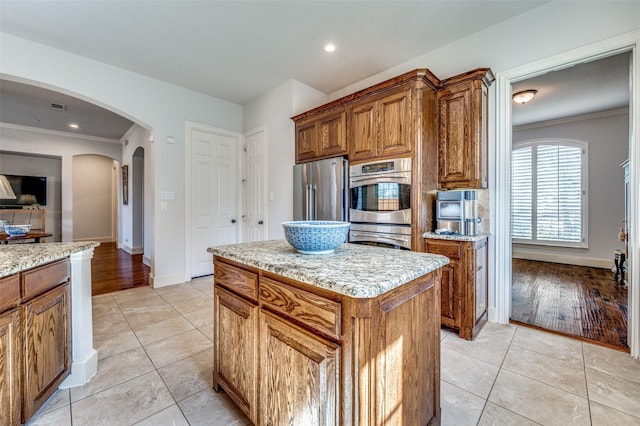 kitchen with light tile patterned floors, brown cabinetry, arched walkways, appliances with stainless steel finishes, and crown molding