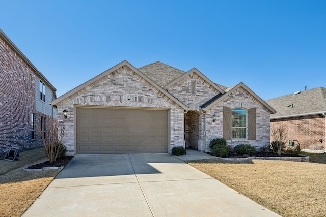 french country style house with a front yard, driveway, an attached garage, a shingled roof, and brick siding