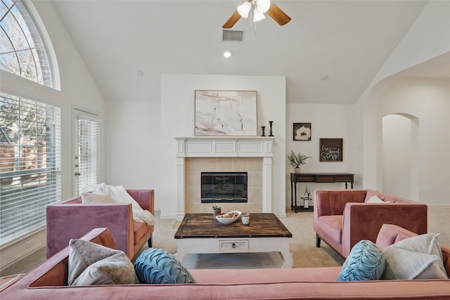 carpeted living room featuring a ceiling fan, visible vents, high vaulted ceiling, arched walkways, and a tiled fireplace