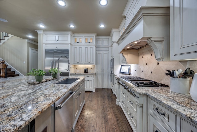 kitchen with a sink, built in appliances, custom exhaust hood, dark wood-style flooring, and light stone countertops