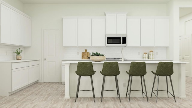 kitchen with white microwave, light countertops, light wood-style flooring, white cabinetry, and a kitchen island with sink