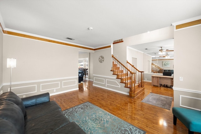 living room featuring a ceiling fan, wood finished floors, crown molding, a decorative wall, and stairs