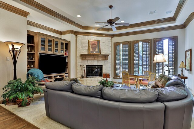 living room with visible vents, a tray ceiling, wood finished floors, a stone fireplace, and crown molding