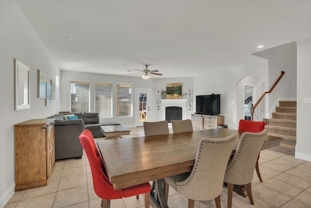 dining room with a fireplace, light tile patterned floors, baseboards, ceiling fan, and stairs