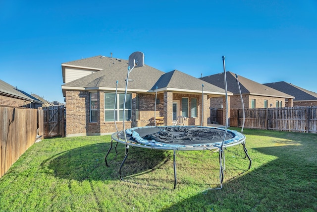 back of house with a trampoline, a fenced backyard, a yard, a shingled roof, and brick siding