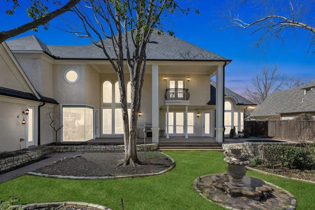 back of property featuring a balcony, fence, a shingled roof, stucco siding, and a lawn