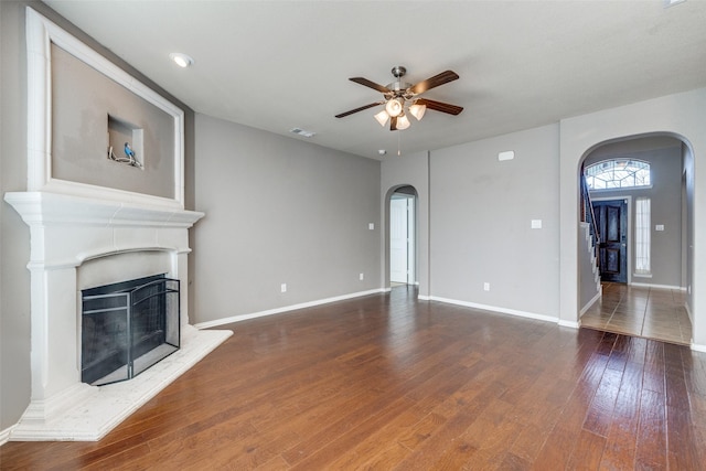 unfurnished living room featuring a ceiling fan, visible vents, a fireplace with raised hearth, arched walkways, and hardwood / wood-style flooring