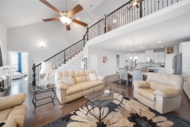 living area featuring visible vents, stairway, ceiling fan with notable chandelier, a towering ceiling, and dark wood-style flooring