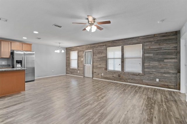 unfurnished living room with visible vents, baseboards, wood walls, light wood-style flooring, and ceiling fan with notable chandelier