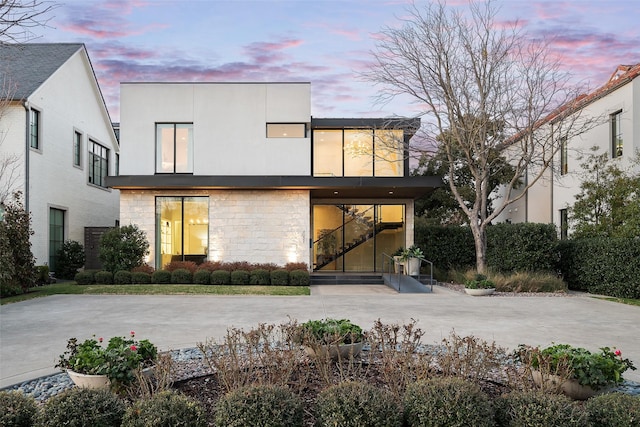 back of house at dusk featuring stone siding, driveway, and stucco siding