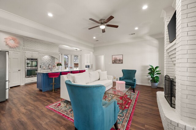 living area with ornamental molding, a ceiling fan, dark wood-style floors, recessed lighting, and a brick fireplace