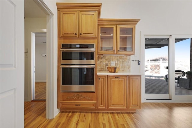 kitchen featuring light wood finished floors, backsplash, glass insert cabinets, light countertops, and stainless steel double oven