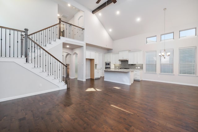 unfurnished living room with beamed ceiling, ceiling fan with notable chandelier, stairway, baseboards, and dark wood-style flooring