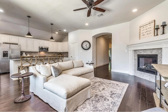 living room with visible vents, dark wood-type flooring, a tiled fireplace, arched walkways, and ceiling fan