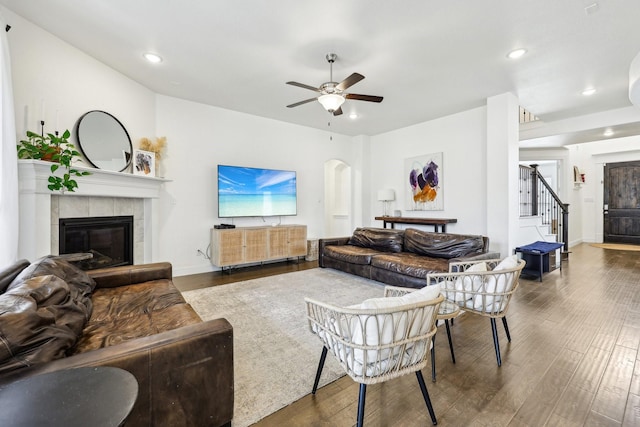 living area featuring stairs, a tile fireplace, wood finished floors, arched walkways, and a ceiling fan