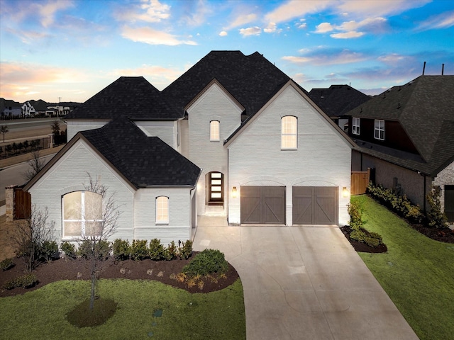 french country inspired facade with roof with shingles, a yard, an attached garage, concrete driveway, and brick siding