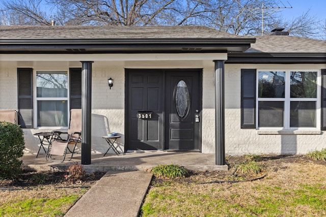 view of exterior entry with covered porch, brick siding, roof with shingles, and a chimney