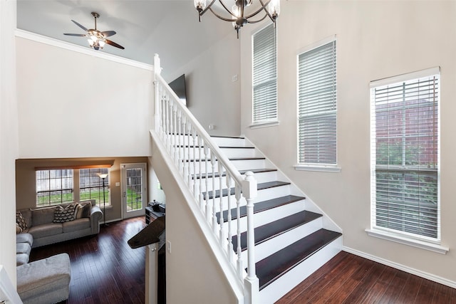 staircase with baseboards, a high ceiling, ornamental molding, wood-type flooring, and ceiling fan with notable chandelier