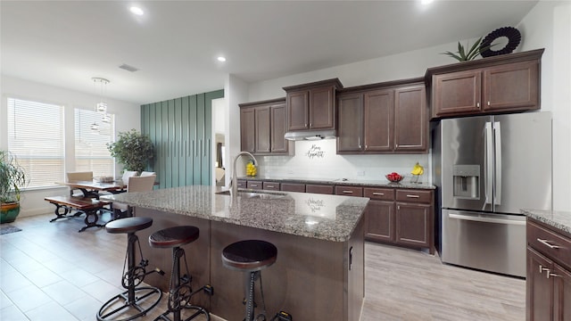 kitchen with a sink, light stone countertops, a kitchen bar, stainless steel fridge, and a kitchen island with sink