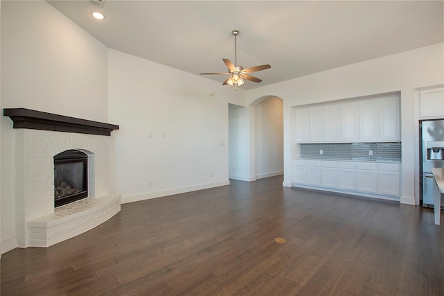 unfurnished living room with a fireplace, arched walkways, dark wood-style flooring, and ceiling fan