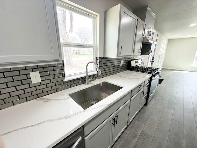 kitchen featuring light stone counters, light wood finished floors, a sink, decorative backsplash, and stainless steel gas range oven