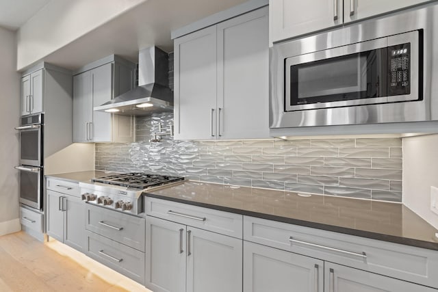 kitchen with light wood-style flooring, dark stone countertops, backsplash, appliances with stainless steel finishes, and wall chimney exhaust hood