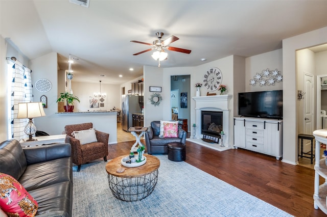 living area featuring visible vents, dark wood-type flooring, lofted ceiling, ceiling fan with notable chandelier, and a glass covered fireplace