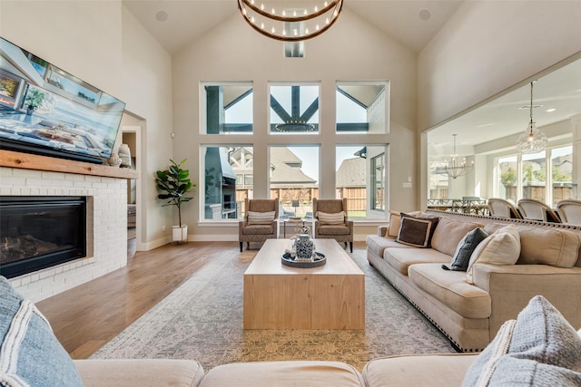 living room with baseboards, high vaulted ceiling, an inviting chandelier, a brick fireplace, and light wood-type flooring