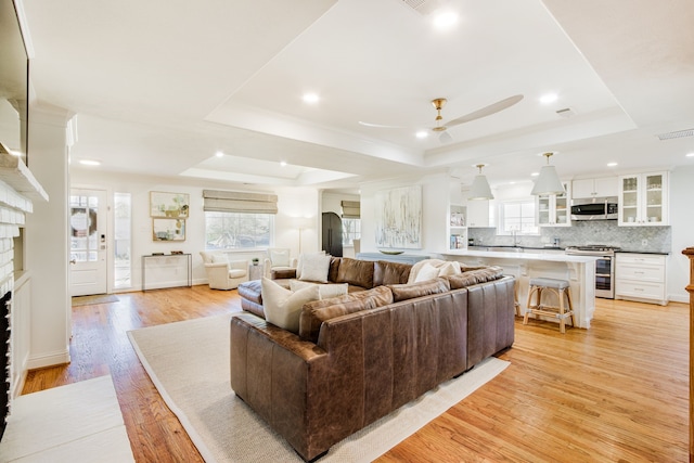 living room with light wood-type flooring, visible vents, a ceiling fan, a raised ceiling, and a brick fireplace